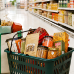 Reusable tote and shopping basket filled with packaged snacks and pantry goods in a supermarket aisle