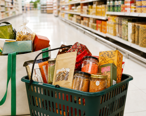 Reusable tote and shopping basket filled with packaged snacks and pantry goods in a supermarket aisle